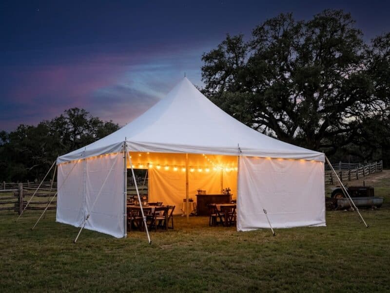 20x20 high peak pole tent rental in Granbury TX with full sidewalls and warm string light glow at night on a ranch property