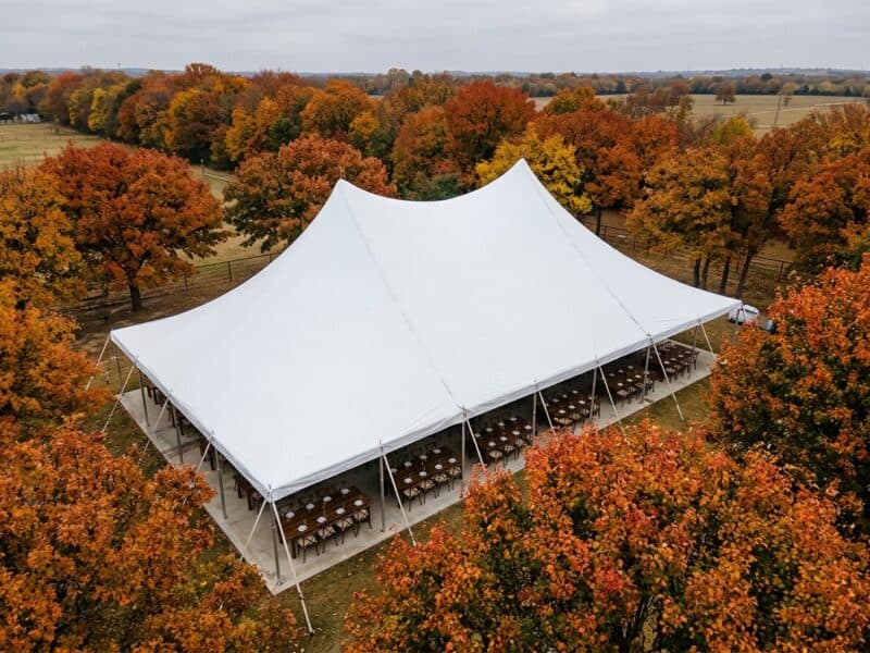 Aerial view of a 30x50 high peak pole tent rental in Alvarado TX with farm tables and cross-back chairs on a concrete pad surrounded by fall foliage