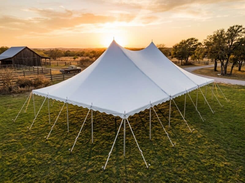 Aerial view of a 30x50 high peak pole tent rental in Granbury TX set up empty on a ranch at sunset with a rustic barn and old pickup truck in the background