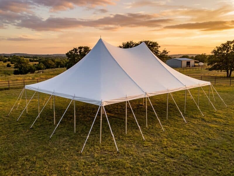 Aerial view of a 30x50 high peak pole tent rental in Godley TX set up empty on a green ranch pasture at sunset with a metal barn in the background