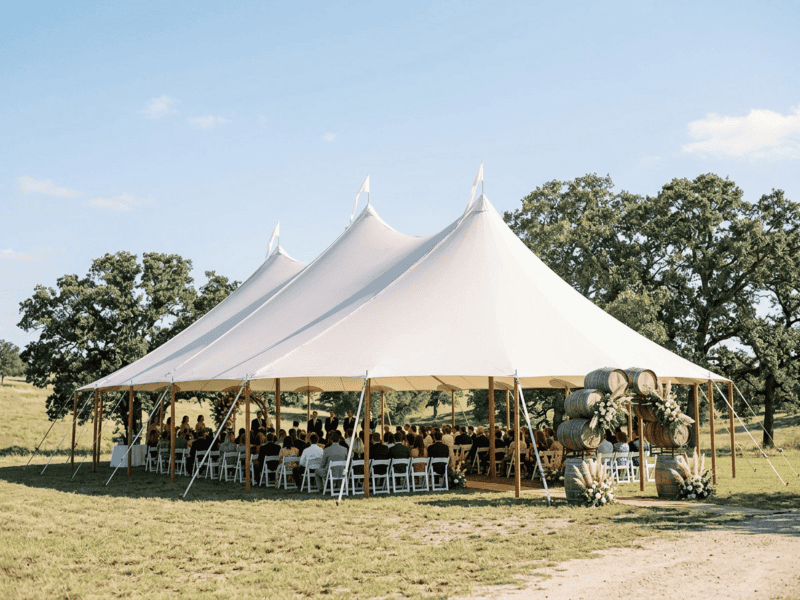 Large three-peak sailcloth tent at open-air wedding ceremony near Burleson TX, stacked wine barrels, guests seated, bright blue sky, wide side angle, Boot & Barrel Event Co.