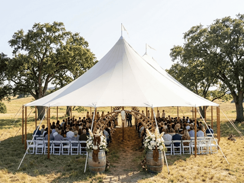 Three-peak sailcloth tent at outdoor wedding ceremony near Weatherford TX, open sides, guests seated in white chairs, floral barrel arch, daytime, Boot & Barrel Event Co.
