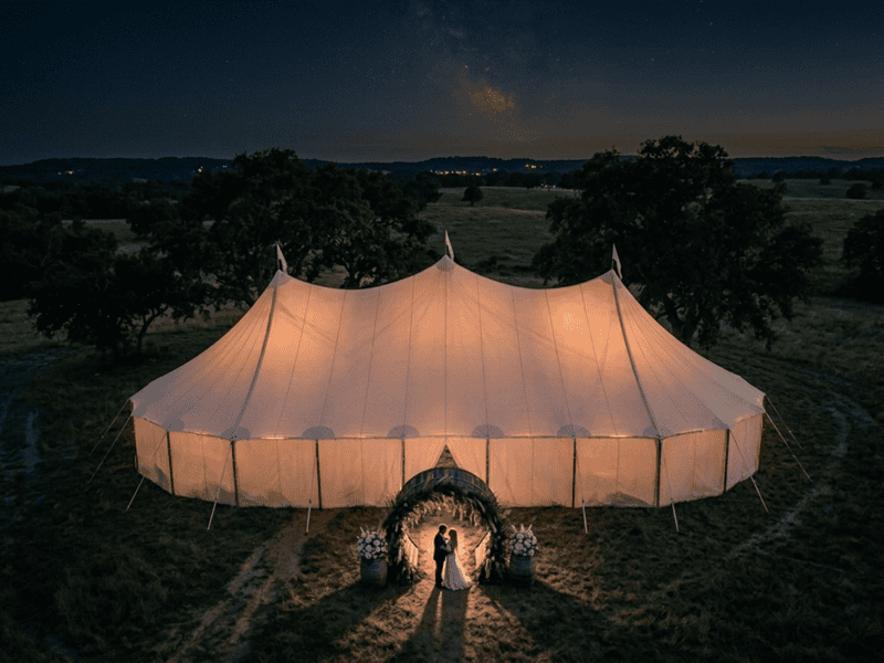 Three-peak sailcloth tent glowing at night with newlywed couple silhouetted in barrel arch entrance, aerial view, Texas ranch, Boot & Barrel Event Co.