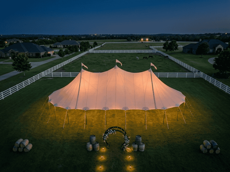 Aerial view of three-peak sailcloth tent glowing at night on manicured estate lawn near Benbrook TX, white ranch fence, horses grazing, barrel arch, Boot & Barrel Event Co.