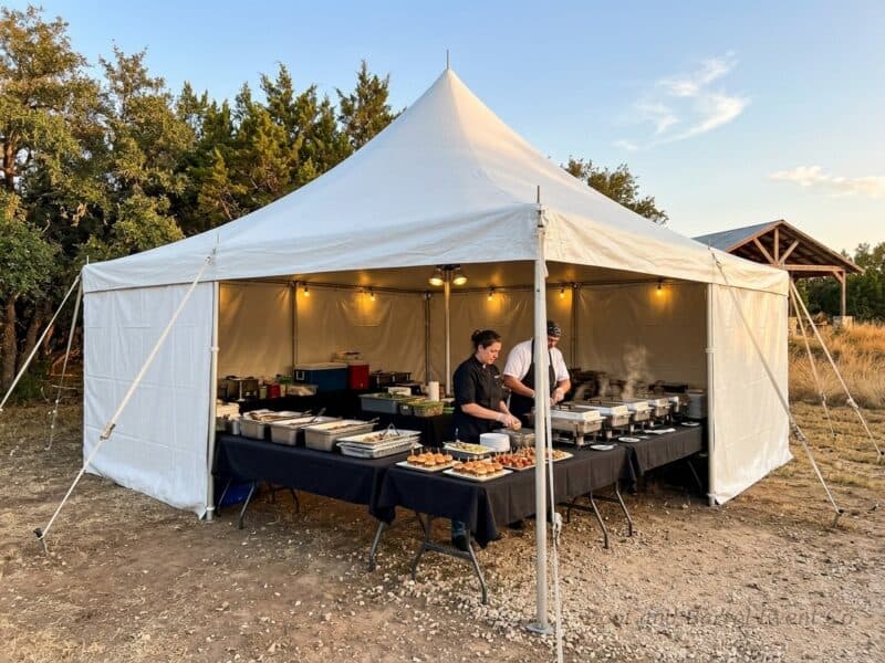 Caterers package tent rental in Granbury TX with catering staff prepping a buffet of sliders and hot dishes under a high peak pole tent at golden hour
