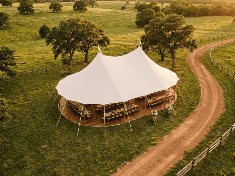 Aerial drone view of 44x63 sailcloth tent with two peaks, farm tables, winding dirt road, split rail fence and Texas ranch landscape