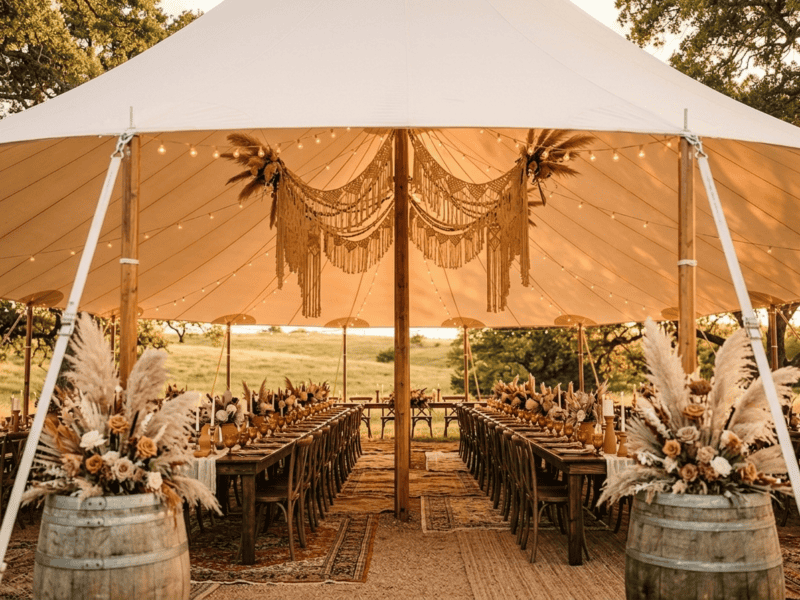 Interior of 44x63 sailcloth tent with boho wedding decor, macrame, pampas grass, farm tables, cross-back chairs and barrel arrangements