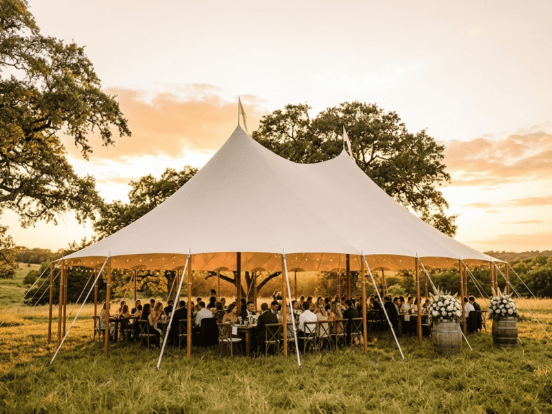 44x63 sailcloth tent wedding reception at golden hour with two peaks, cross-back chairs, barrel decor and oak trees in Texas pasture