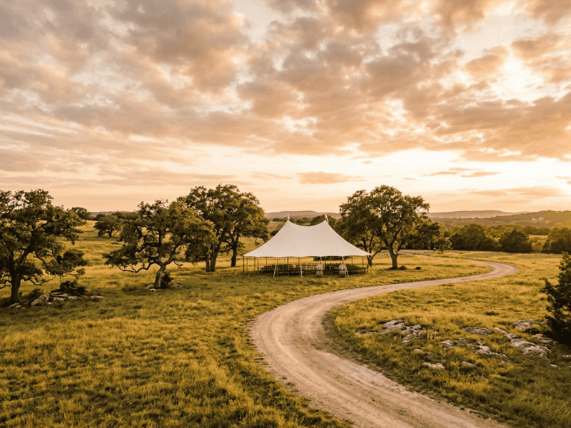 Wide landscape view of 44x63 sailcloth tent with two peaks on winding dirt road in Texas Hill Country at golden hour