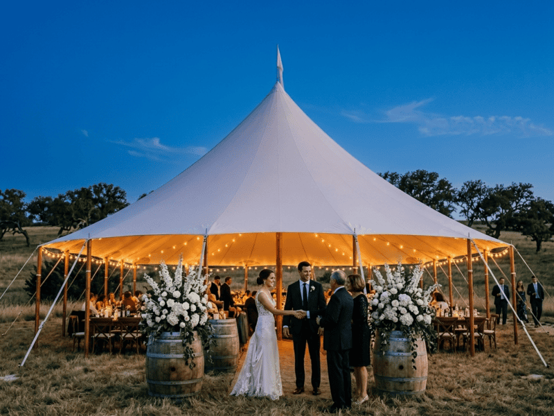Sailcloth tent wedding at blue hour with bride and groom at barrel entrance, white floral arrangements and string lights glowing inside