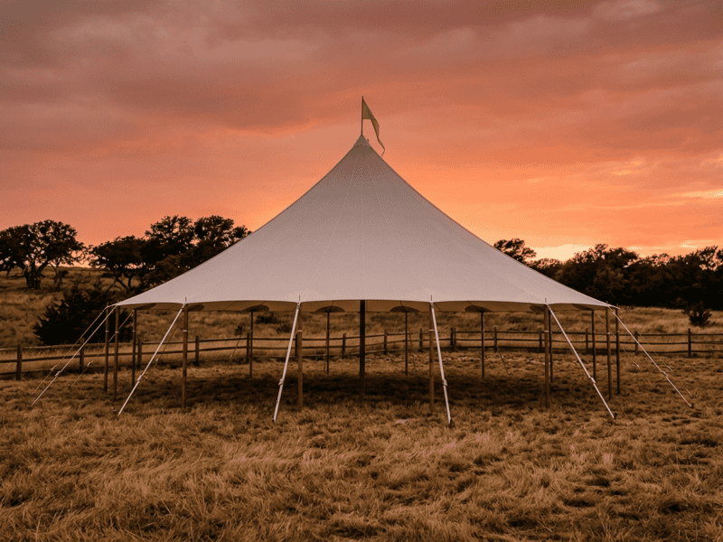 Oval sailcloth tent at dramatic orange sunset with wooden poles, split rail fence and dry Texas pasture — no people
