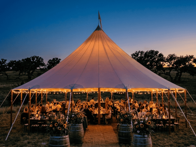 Oval sailcloth tent wedding reception at dusk with string lights, sunflower barrel decor and guests dining in Texas Hill Country