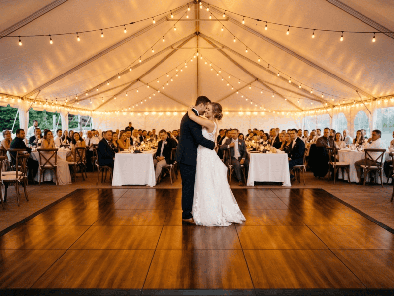 Bride and groom first dance on fruitwood portable dance floor rental under white tent with Edison string lights — Boot & Barrel Event Co. Mansfield TX